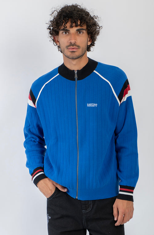 Chaqueta de punto azul con cremallera, cuello bomber y detalles deportivos en blanco, negro y rojo.