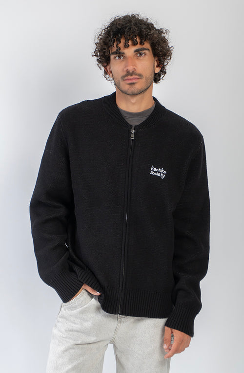 Jersey unisex de punto negro con cremallera, bordado Kaotiko Society en el pecho y gráfico jacquard de flores azules con mensaje Bad Romance en la espalda.